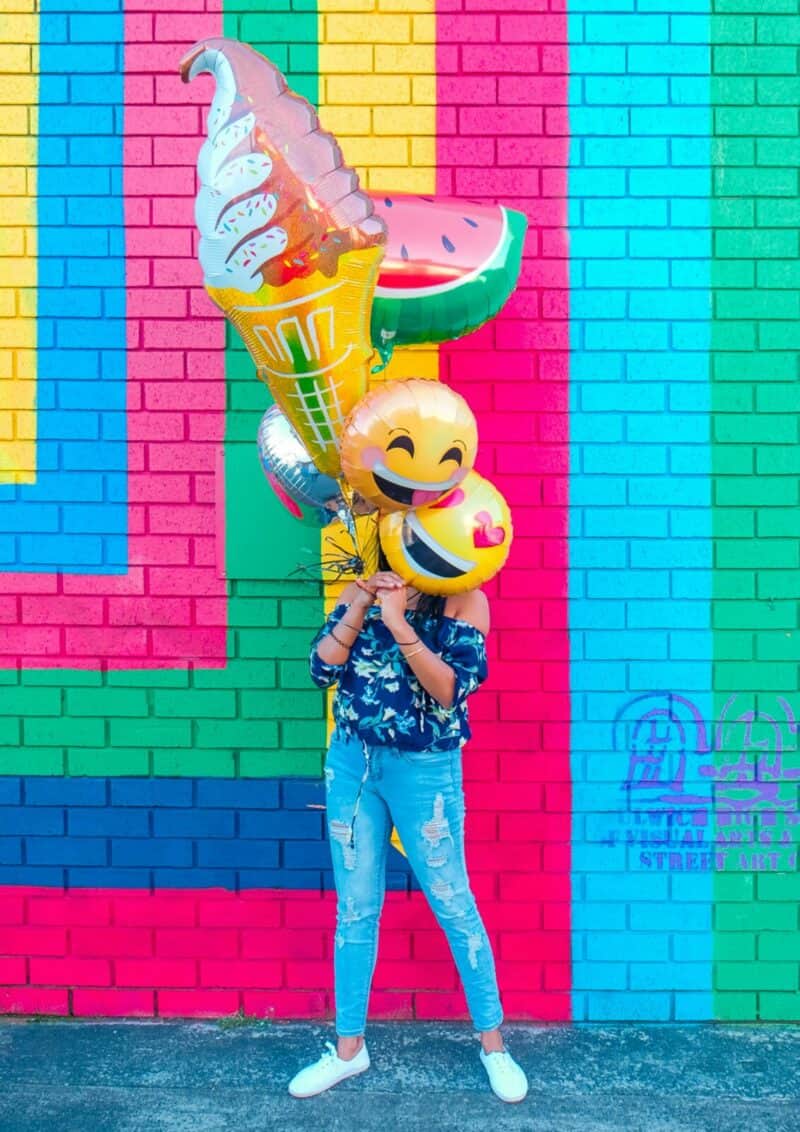 Girl holding emoji balloons in front of her face standing in front of colorful stripped wall Photo by Lidya Nada on Unsplash